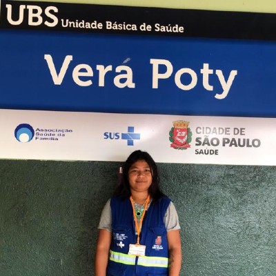 Foto de Clarice Honório.  Em pé, diante da fachada da UBS onde trabalha, Clarice está sorrindo diante da placa azul e branca com o nome Vera Poty em letras grandes. Ela tem cabelos negros lisos e usa colete azul, um crachá pendurado com fita laranja no pescoço e usa pulseiras de artesanato nos dois braços